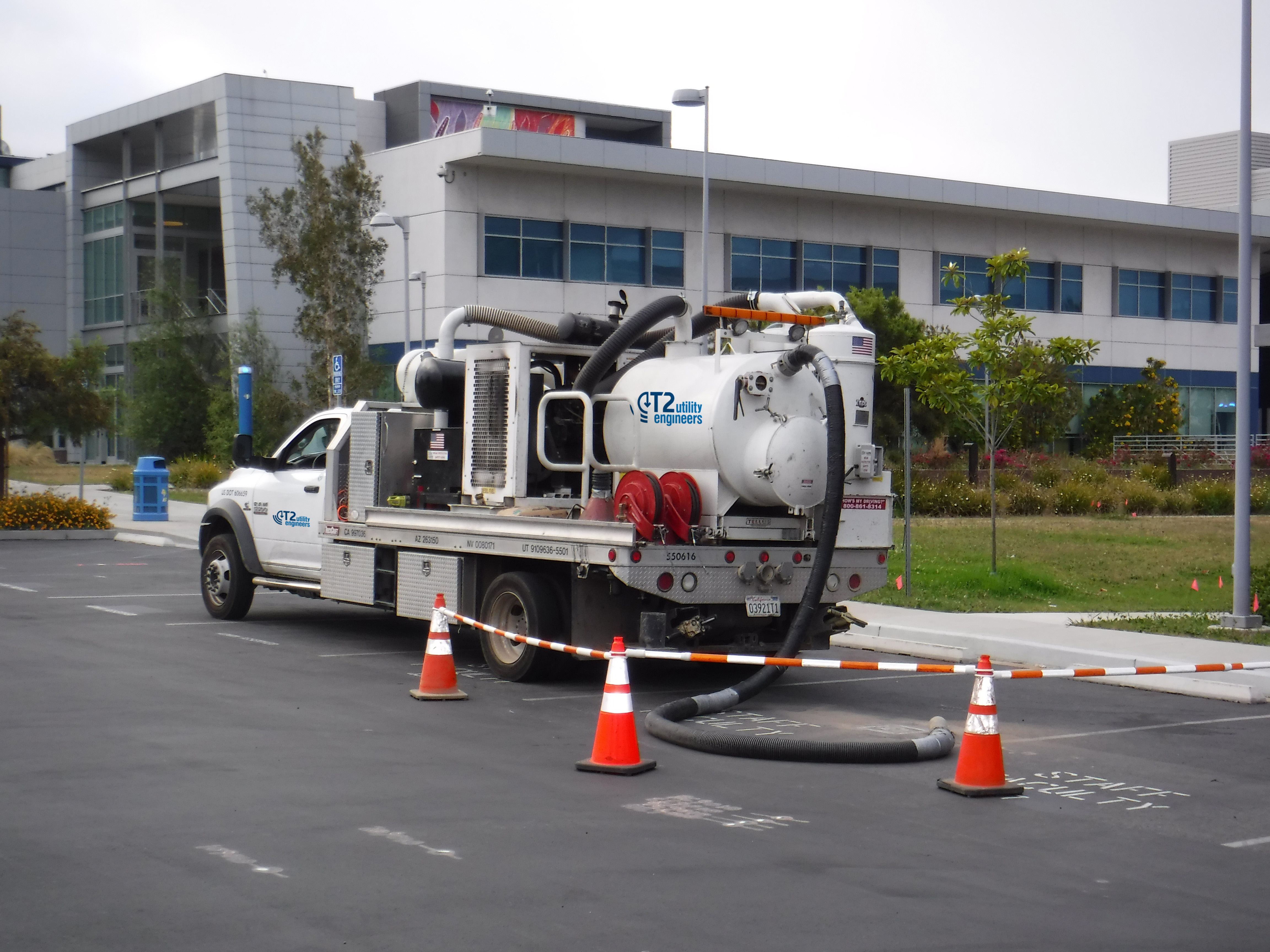 Utility service truck with a large vacuum tank and hoses parked outside a school facility, surrounded by traffic cones.