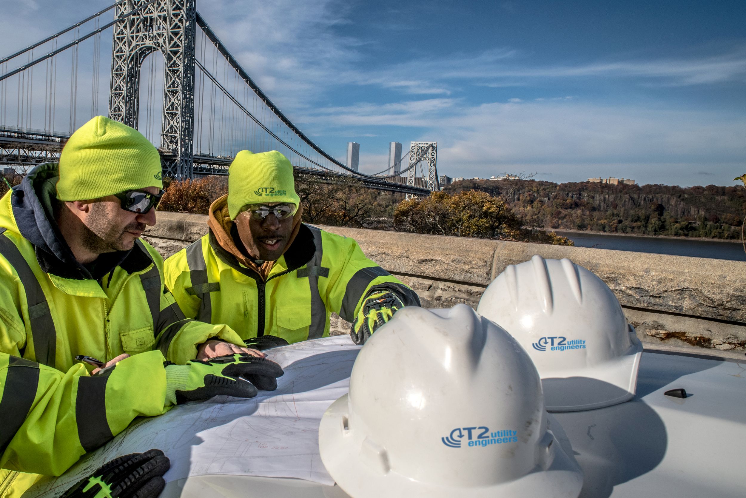 Construction workers in high-visibility jackets and neon beanies reviewing plans near the George Washington Bridge, with white hard hats on a vehicle in the foreground.