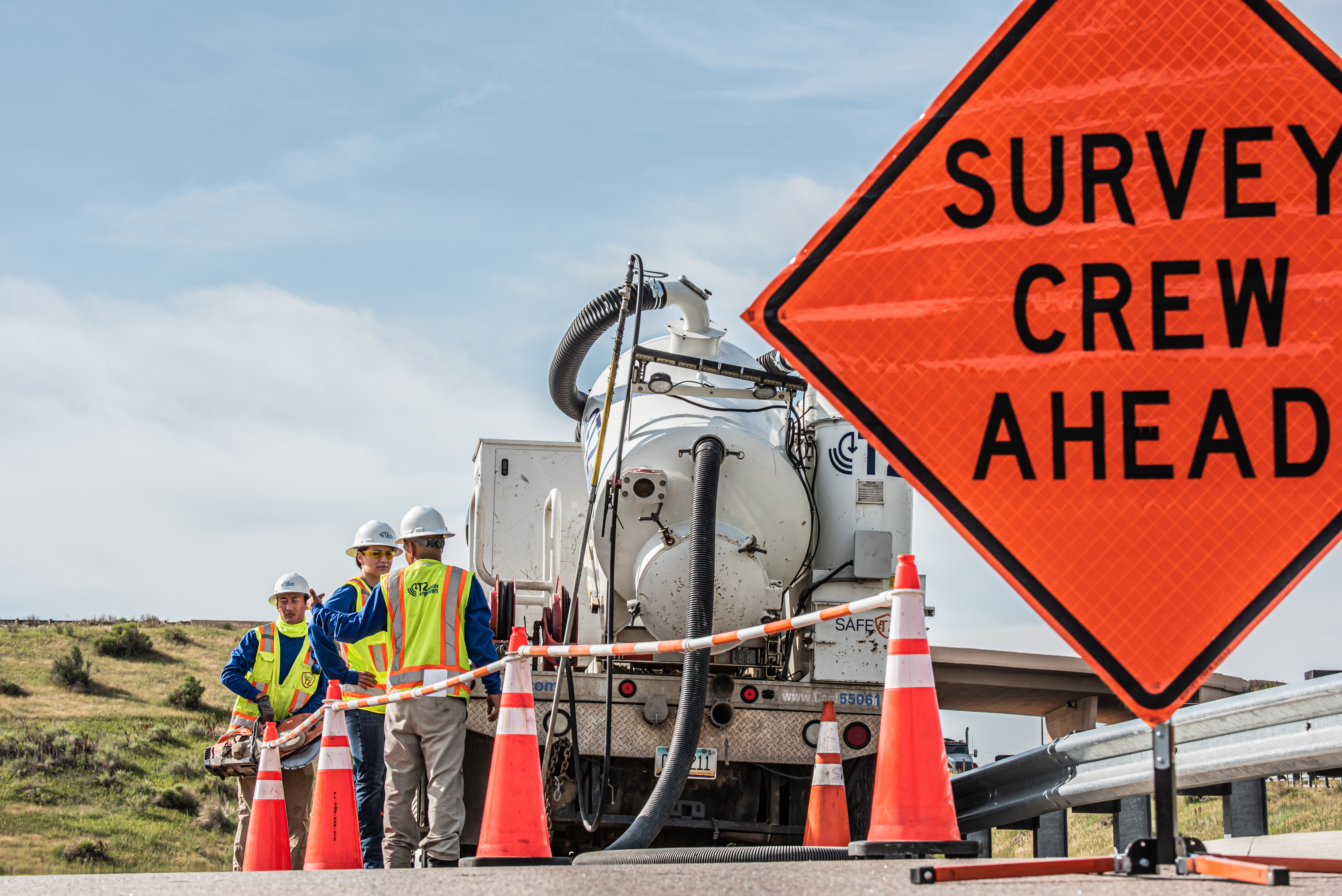 Road work scene with survey crew ahead sign, traffic cones, and workers beside a utility truck on a rural roadside.