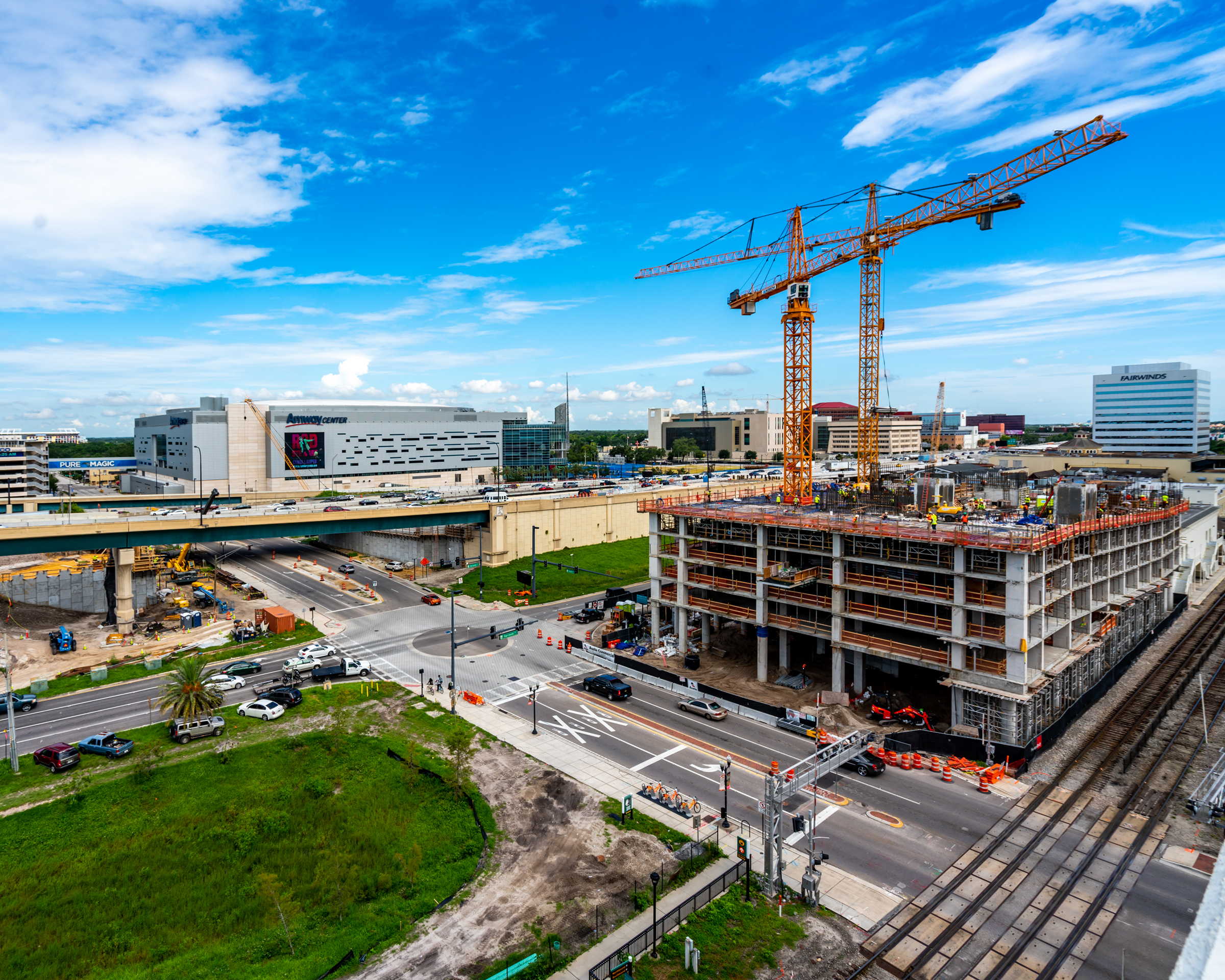 Urban construction site with two tower cranes, a partially built high-rise, and surrounding city roads near a large arena under a blue sky.