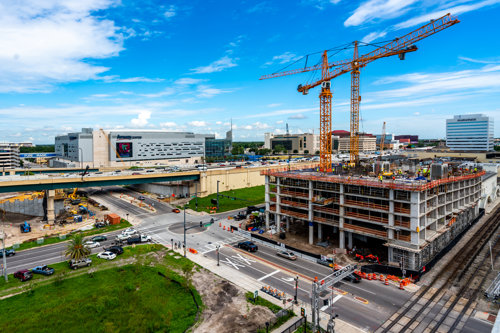 Urban construction site with two tower cranes, a partially built high-rise, and surrounding city roads near a large arena under a blue sky.