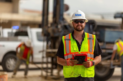 Construction worker in a hard hat and high-visibility safety vest holding a tablet at a work site, with heavy equipment and other workers in the background.