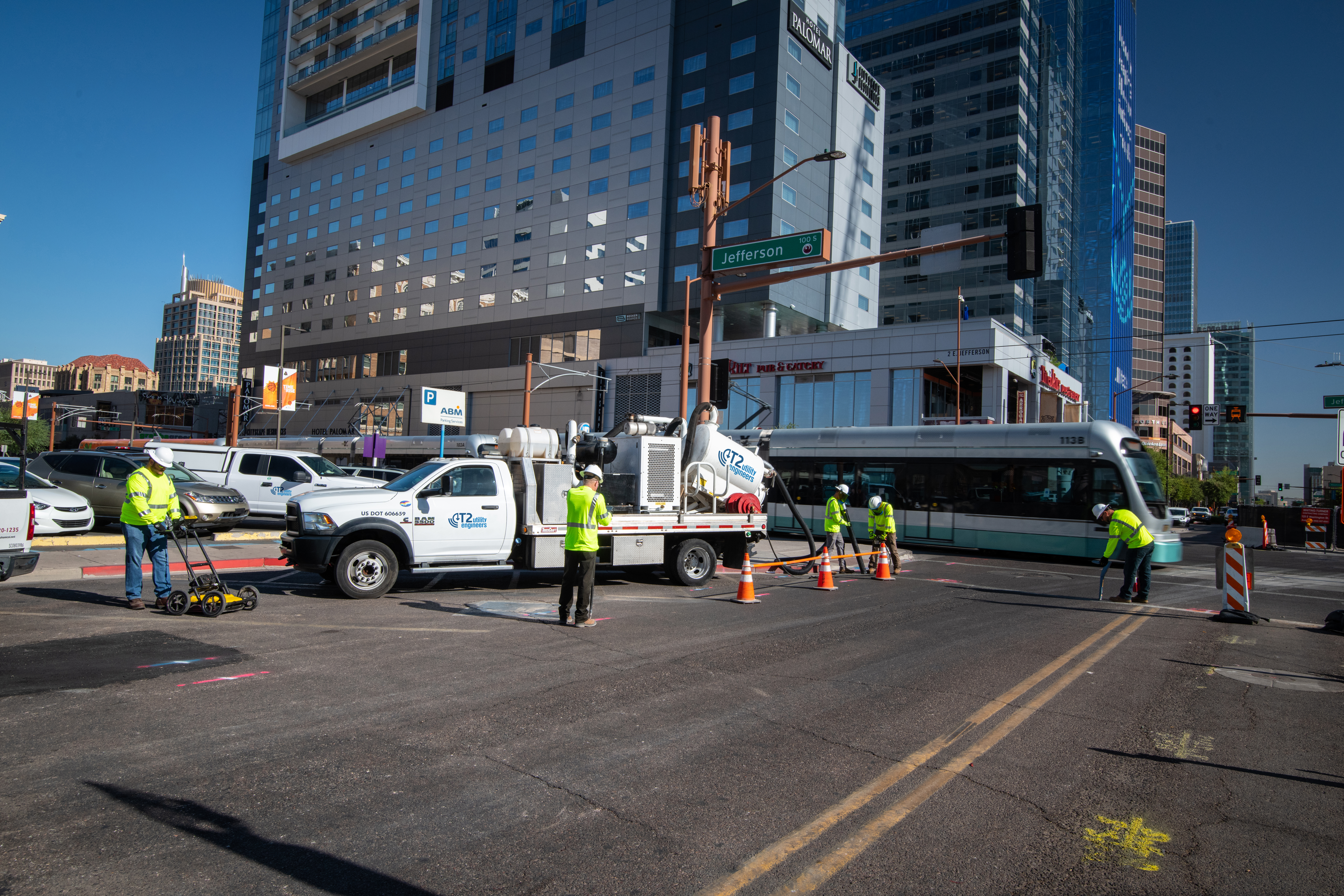 Kiwa USA employees in reflective vests repairing pavement at a downtown street intersection with a city bus passing by and tall buildings in the background.