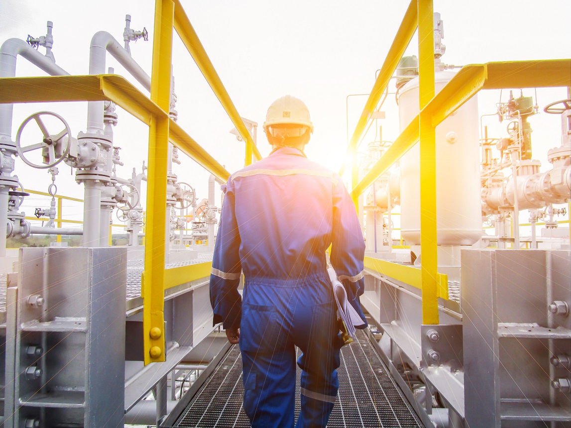 Industrial worker in blue coveralls and a hard hat walking through an oil and gas facility with yellow railings and pipes.