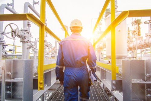 A technician in blue coveralls and a hard hat is walking through an industrial plant with pipes, valves and yellow railings